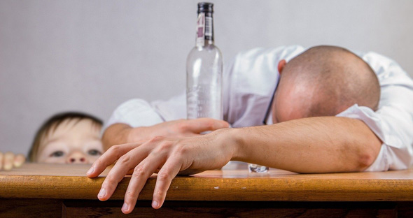 Mann mit leerer Flasche schläft auf Tischplatte, hinter der ein Kind hervorlugt