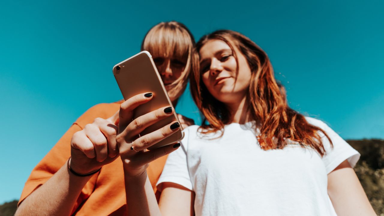Zwei junge Frauen stehen Seite an Seite und lesen gemeinsam die Social-Media-Beiträge, Nachrichten und Mitteilungen auf einem Mobiltelefon. Aufnahme von unten vor dem Sommerhimmel.