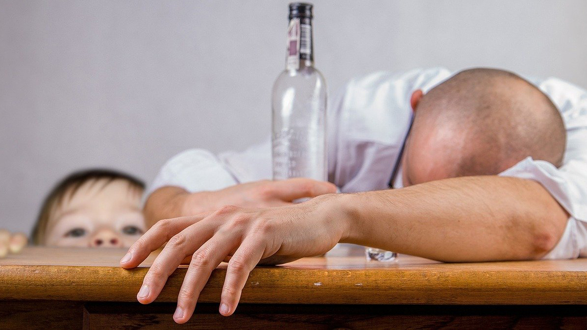 Kind lugt über Tischkante,; auf dem Tisch schlafender Mann mit leerer Flasche