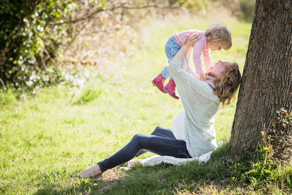 Mutter hebt Kind über ihren Kopf, während sie in einem Park an einem Baum sitzt