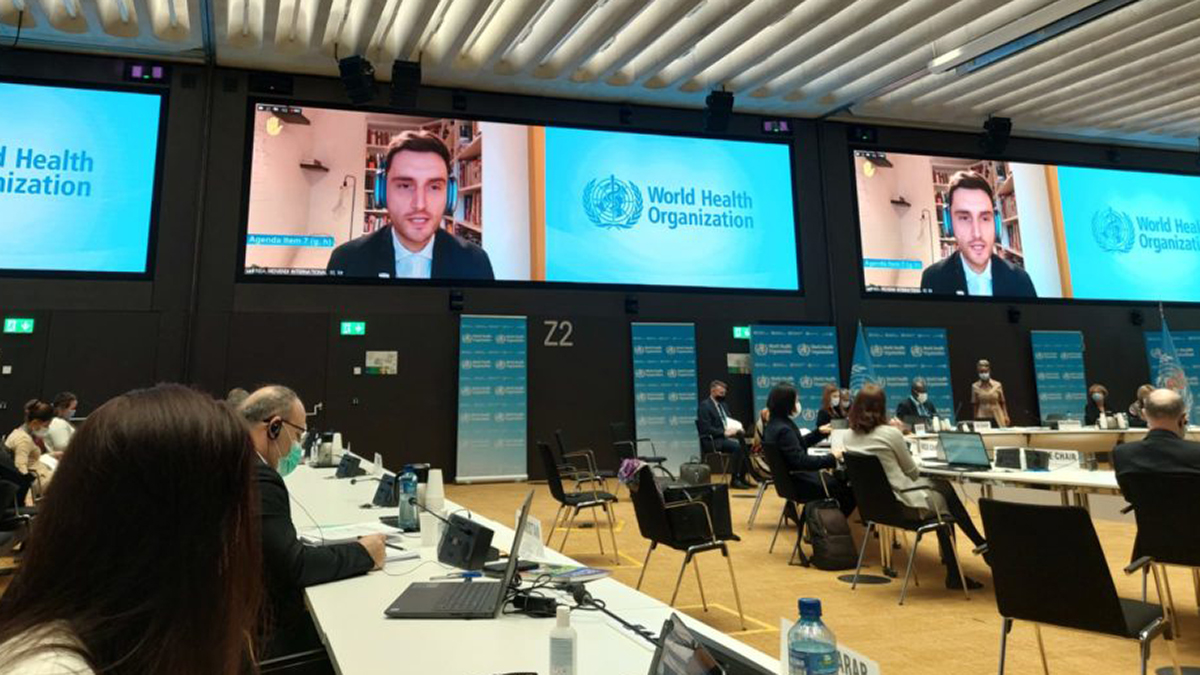 Maik Dünnbier auf Konferenzbildschirmen bei der Abgabe der Erklärung von Movendi International während EB150 im Konferenzsaal