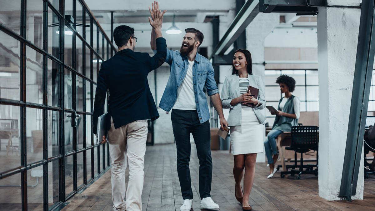 Zwei Männer klatschen bei Begnung im Büroflur "High Five". Begleitende Frau schaut zu.