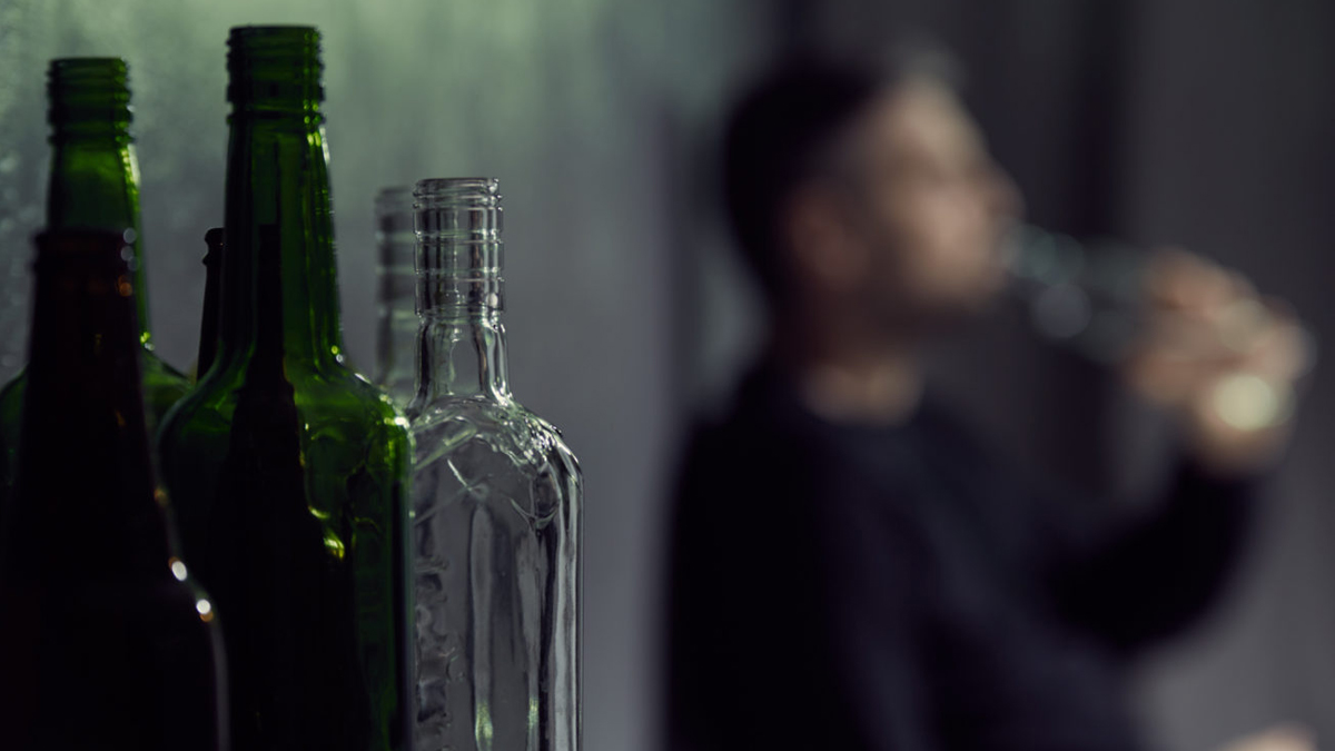 Leere Alkoholflaschen im Vordergrund, im Hintergrund verschwommen erkennbar ein trinkender Mann