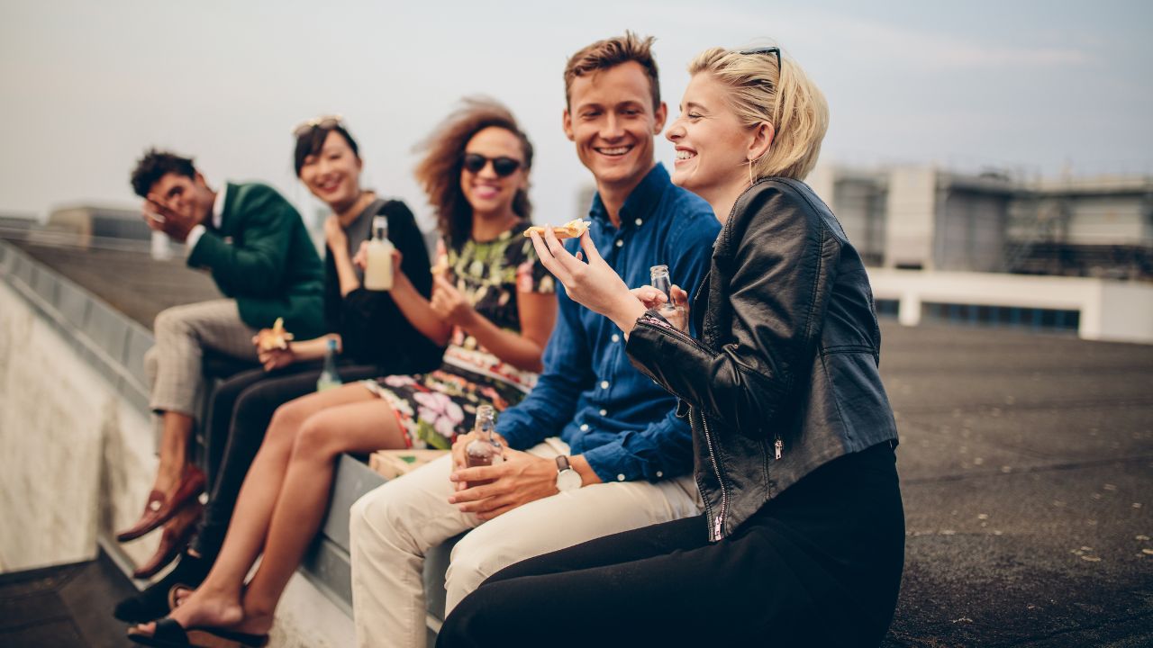 Fünf junge Erwachsene sitzen mit Partyhäppchen und Getränkeflaschen auf der Kante eines Daches.