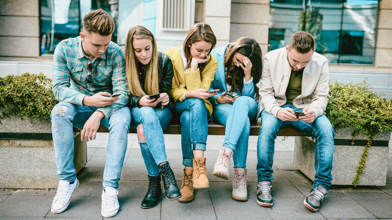 Fünf Jugendliche sitzen nebeneinander auf einer Bank und betrachten ihr jeweiliges Smartphone.
