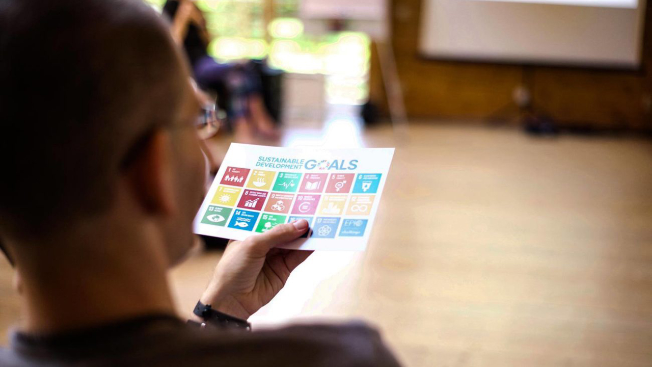 Blick über die Schulter eines Mannes, der auf einer Couch sitzt und eine Karte mit den Logos der Nachhaltigen Entwicklungsziele betrachtet.