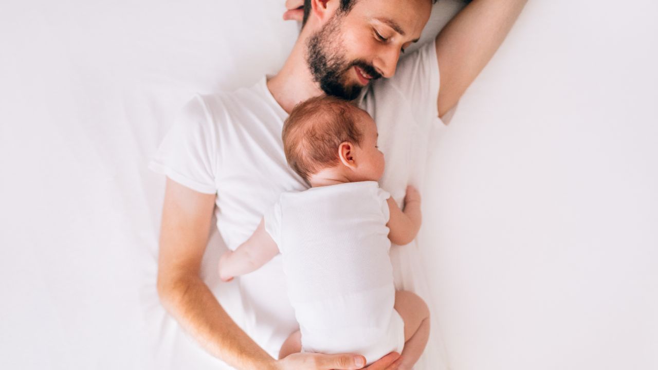 Vater liegt mit Baby auf seinem Bauch auf einer weißen Matratze.