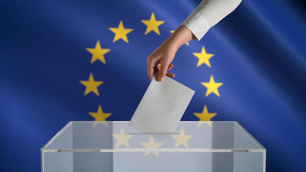 Eine Hand steckt einen Wahlumschlag in eine transparente Wahlurne. Im Hintergrund die Europa-Flagge.