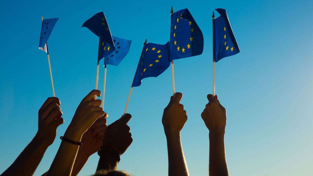 Sechs Hände winken mit Europa-Fähnchen vor blauem Himmel.