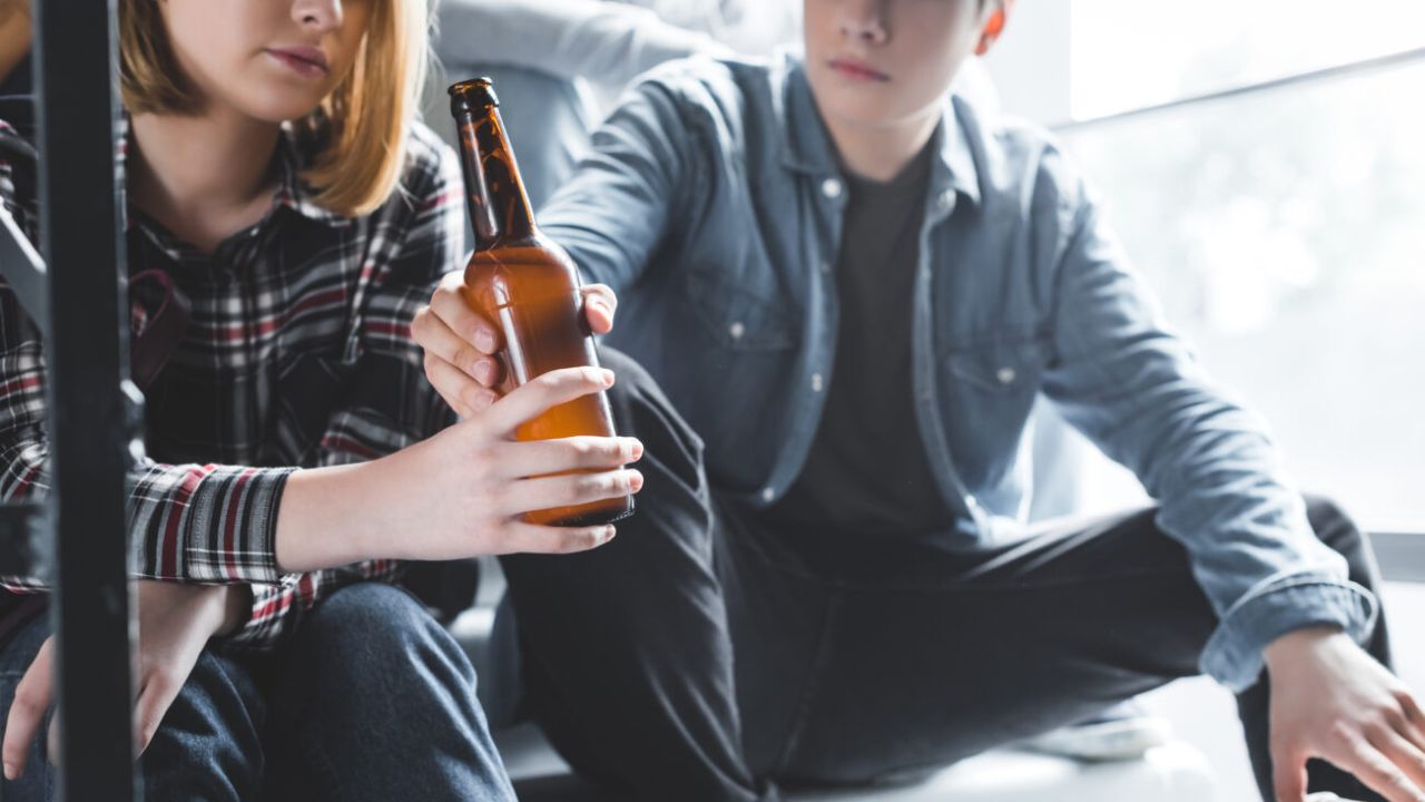 Ausschnitt eines Jungen, der einem Mädchen eine Glasflasche mit Bier gibt.