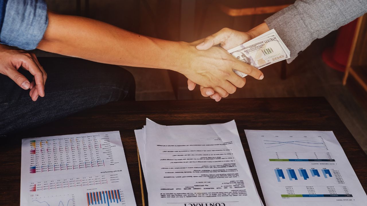 Händedruck zweier Männer über Geschäftspapieren, bei dem Dollarnoten übergeben werden.