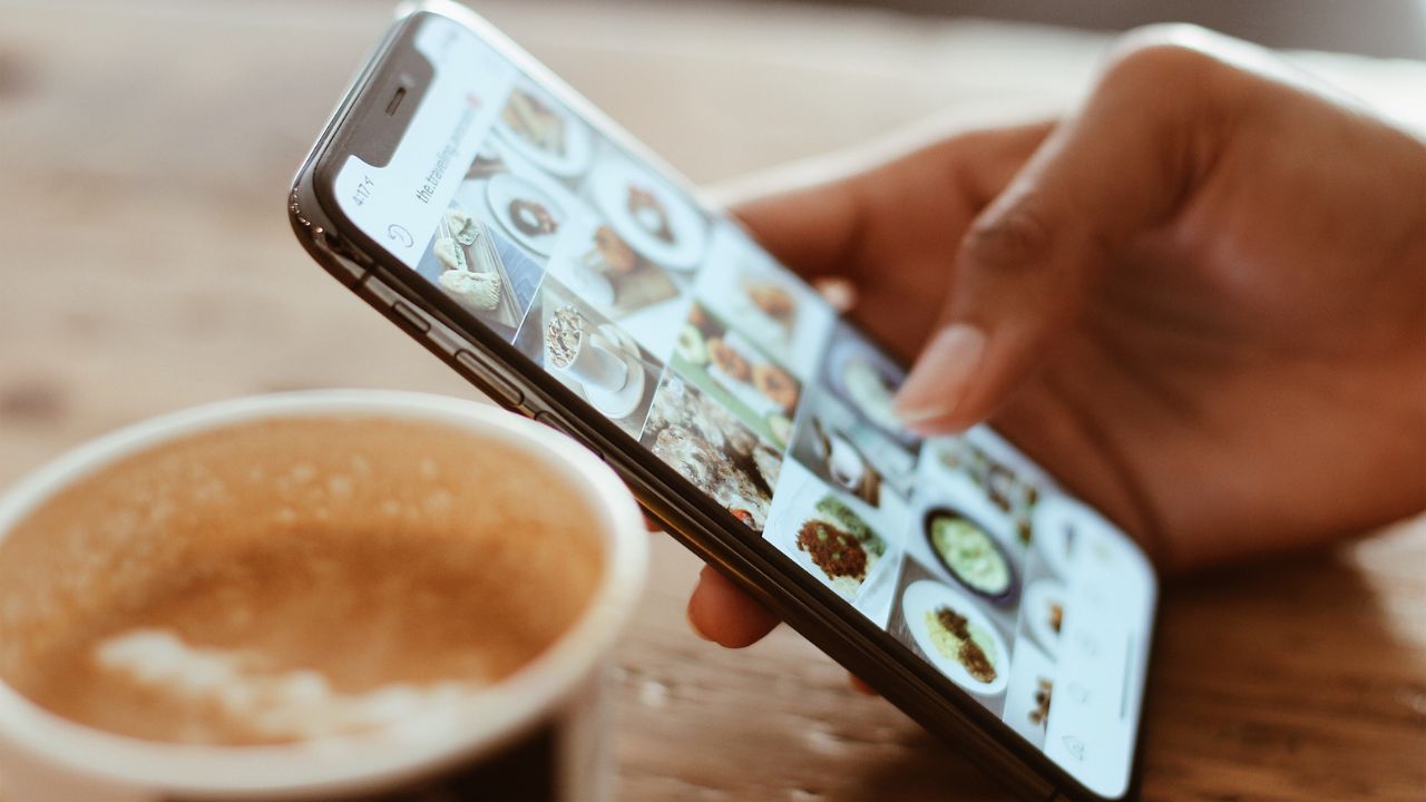 Hand hält ein eingeschaltetes Smartphone neben einem Kaffeebecher auf einem Tisch.