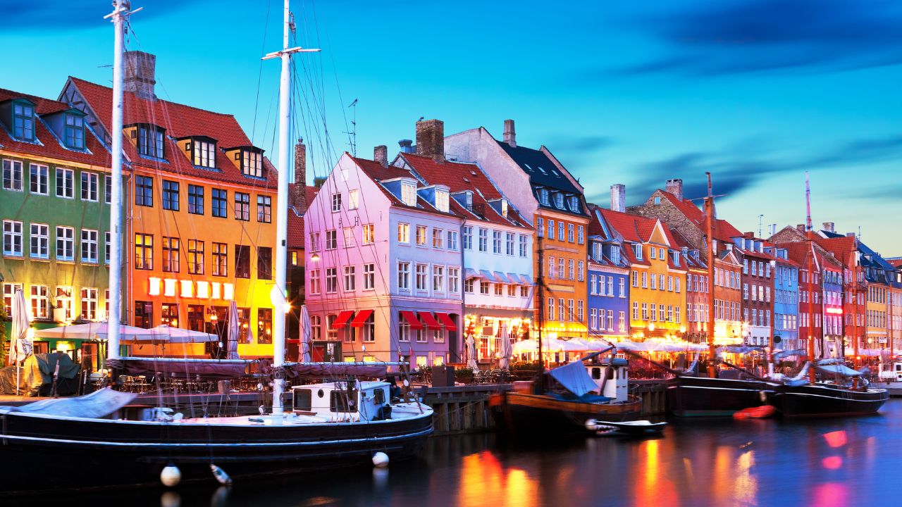 Bunte Häuser am Hafen von Nyhavn in Kopenhagen bei Abenddämmerung mit beleuchteten Restaurants und Segelbooten im Wasser.