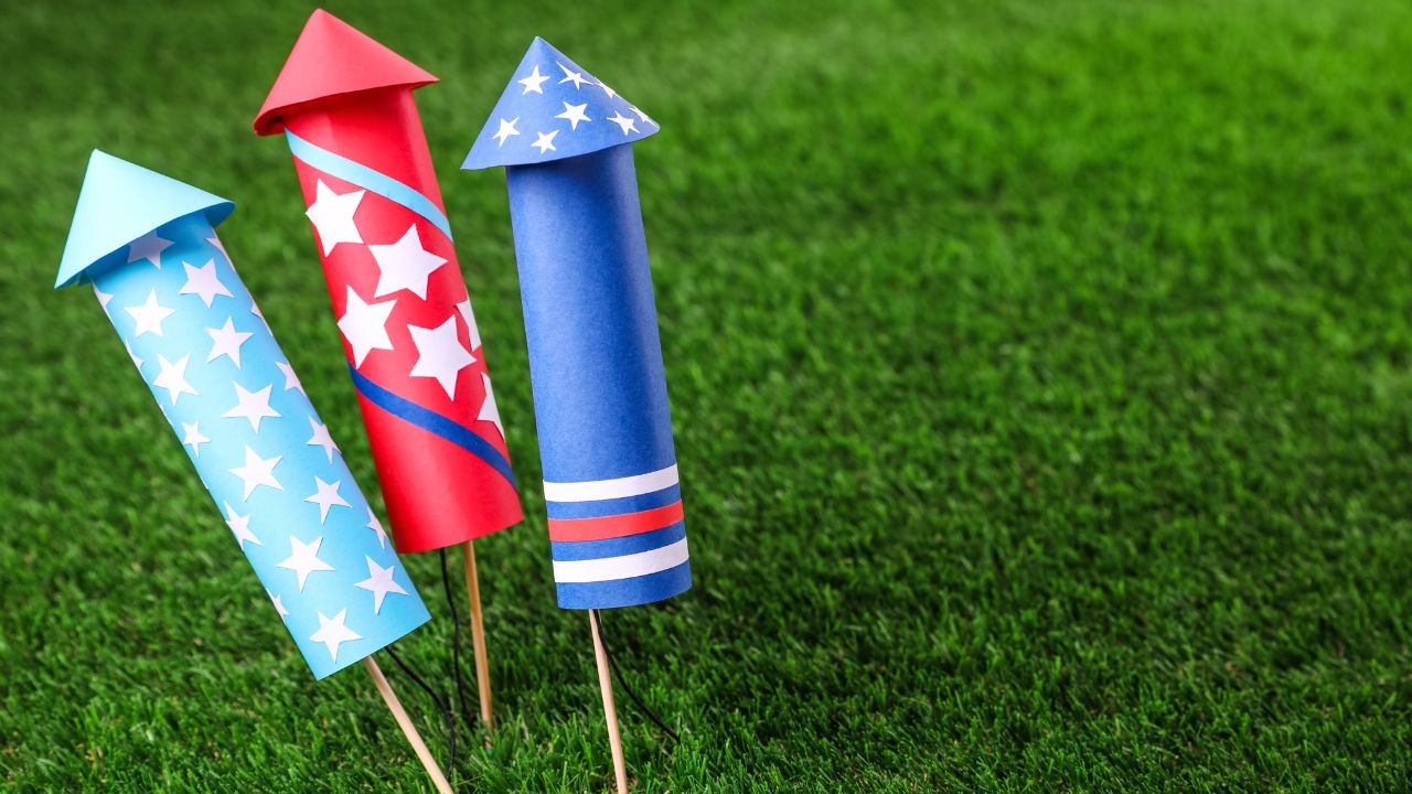 Drei bunte Feuerwerksraketen mit Sternenmuster in Rot, Blau und Weiß stehen auf grünem Rasen.