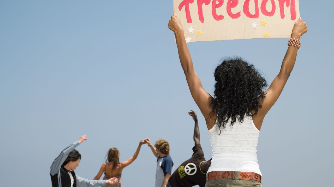 Rückenansicht einer jungen Frau mit dunklen Locken, die ein handgemaltes Schild mit dem roten Schriftzug 'Freedom' und kleinen Sternen hochhält. Im Hintergrund sind weitere junge Menschen zu sehen, die mit erhobenen Armen tanzen oder jubeln, vor einem klaren blauen Himmel.