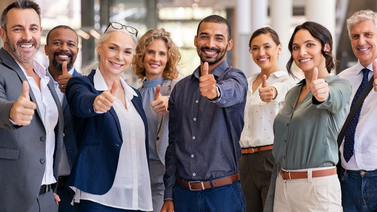 Eine diverse Gruppe von acht geschäftlich gekleideten Personen unterschiedlichen Alters und verschiedener Ethnien steht in einem hellen Bürogebäude nebeneinander. Alle lächeln freundlich in die Kamera und zeigen mit der rechten Hand eine 'Daumen hoch'-Geste als Zeichen von Zustimmung und Erfolg.