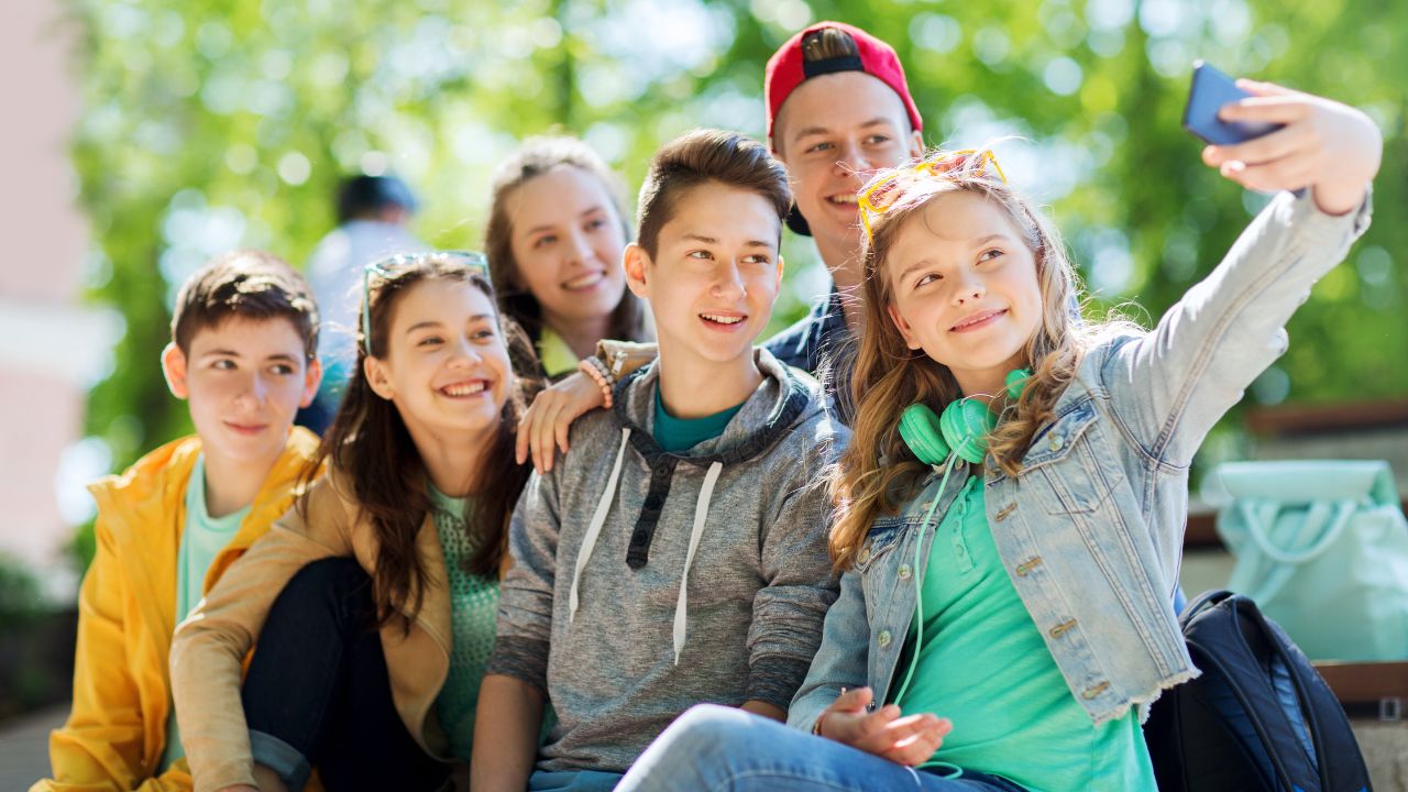 Eine Gruppe fröhlicher Jugendlicher sitzt im Freien zusammen und macht ein Selfie. Alle lächeln und wirken entspannt. Im Hintergrund sind grüne Bäume und Sonnenlicht zu sehen, was eine freundliche, sommerliche Atmosphäre schafft.