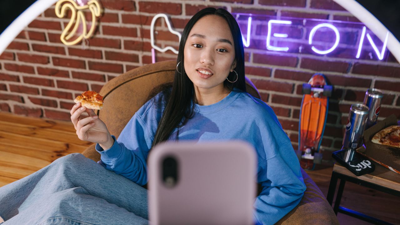 Eine junge Frau sitzt entspannt in einem braunen Sitzsack und hält ein Stück Pizza in der rechten Hand. Sie trägt ein blaues Sweatshirt und Jeans und blickt direkt in die Kamera eines Smartphones im Vordergrund, das wahrscheinlich für ein Video oder Selfie genutzt wird. Der Hintergrund zeigt eine Ziegelsteinwand mit lila Neonschrift, ein Skateboard und Getränkedosen auf einem Tisch.