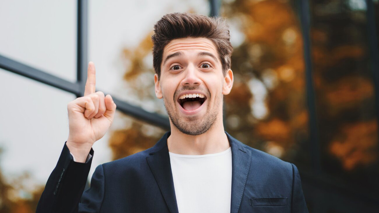 Ein junger Mann in einem blauen Anzug zeigt mit dem Zeigefinger nach oben und lächelt breit, als würde er eine plötzliche Idee oder einen Einfall haben. Sein Gesichtsausdruck strahlt Freude und Begeisterung aus.