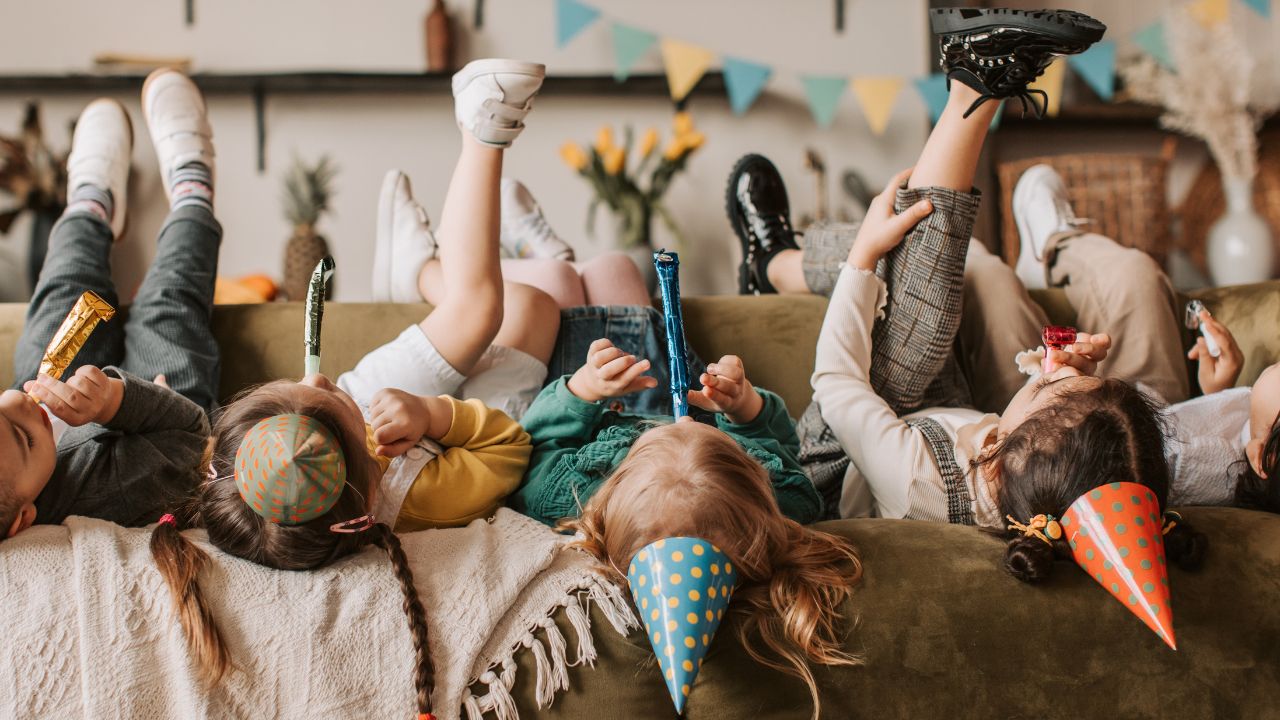 Eine Gruppe von fünf Kindern feiert liegend auf einer grünen Couch, mit Partyhüten und Partytröten, während ihre Beine und Füße nach oben in die Luft ragen.