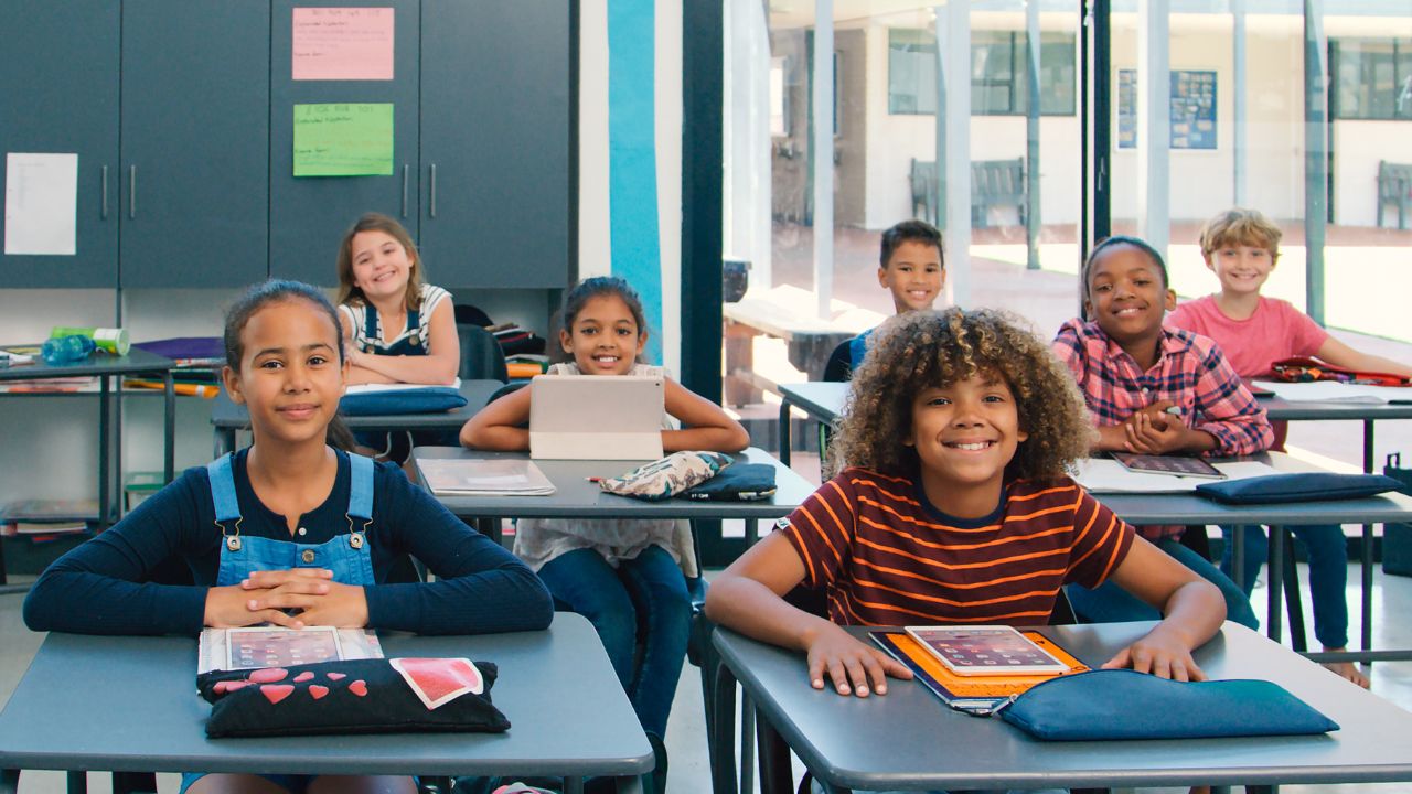 Sieben diverse Grundschulkinder unterschiedlicher Herkunft sitzen in drei Reihen an Schulpulten in einem offenen hellen Klassenzimmer. Alle Kinder schauen freundlich und lächelnd direkt in die Kamera. Auf den Pulten liegen Tabletts und Federmäppchen.