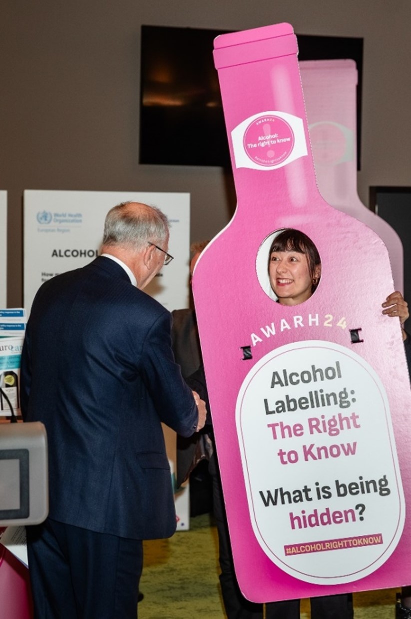 Der Europaabgeordnete Vytenis Povilas Andriukaitis schüttelt dem Kampagnen-Maskottchen die Hand, das die Kernbotschaft 'Alkoholkennzeichnung: Das Recht auf Information. Was wird verheimlicht?' auf einer Flaschen-Verkleidung trägt.