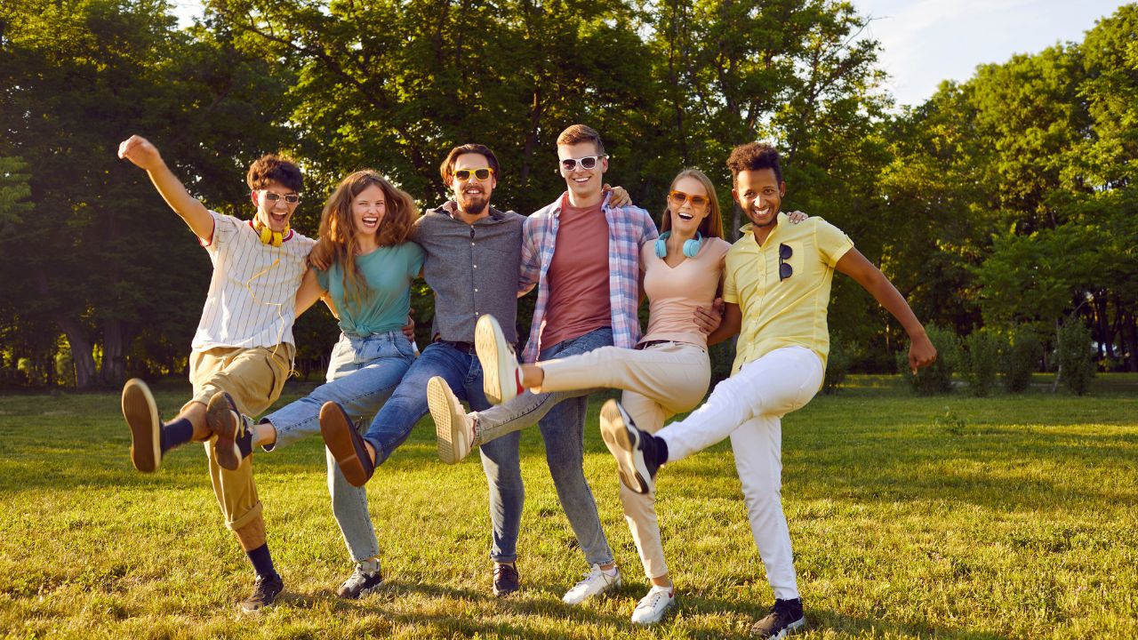 Eine vielfältige Gruppe von sechs jungen Erwachsenen steht Arm in Arm auf einer sonnigen Wiese. Sie lachen ausgelassen und kicken synchron ein Bein in die Luft, im Hintergrund sind grüne Bäume zu sehen.