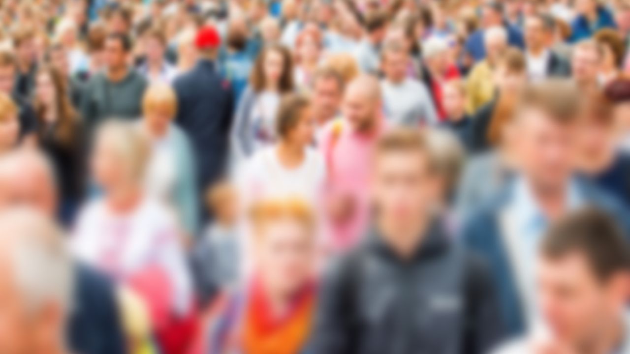 Verschwommenes Bild einer großen Menschenmenge bei einer Veranstaltung. Viele Personen stehen dicht beieinander, erkennbar an verschiedenfarbiger Kleidung in Rot, Pink, Blau und anderen Farben. Die bewusste Unschärfe lässt keine individuellen Gesichter erkennen.