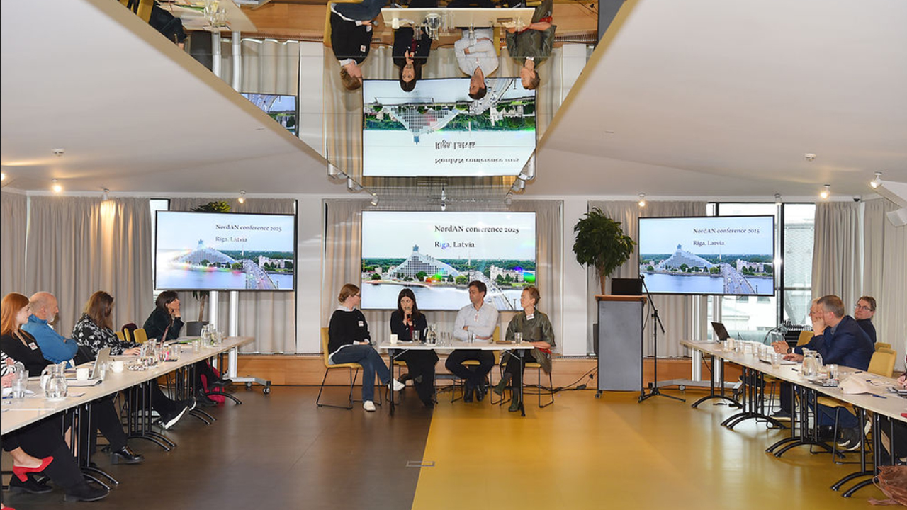 Ein Konferenzraum während einer Präsentation mit dem Titel 'NordAN Conference 2025' in Riga, Lettland. Im Vordergrund sitzt eine fünfköpfige Gruppe an einem Podiumstisch, während das Publikum an langen Tafeln Platz genommen hat. Drei große Bildschirme zeigen die Präsentation. Eine markante, spiegelnde Deckenkonstruktion reflektiert die Szene darunter.
