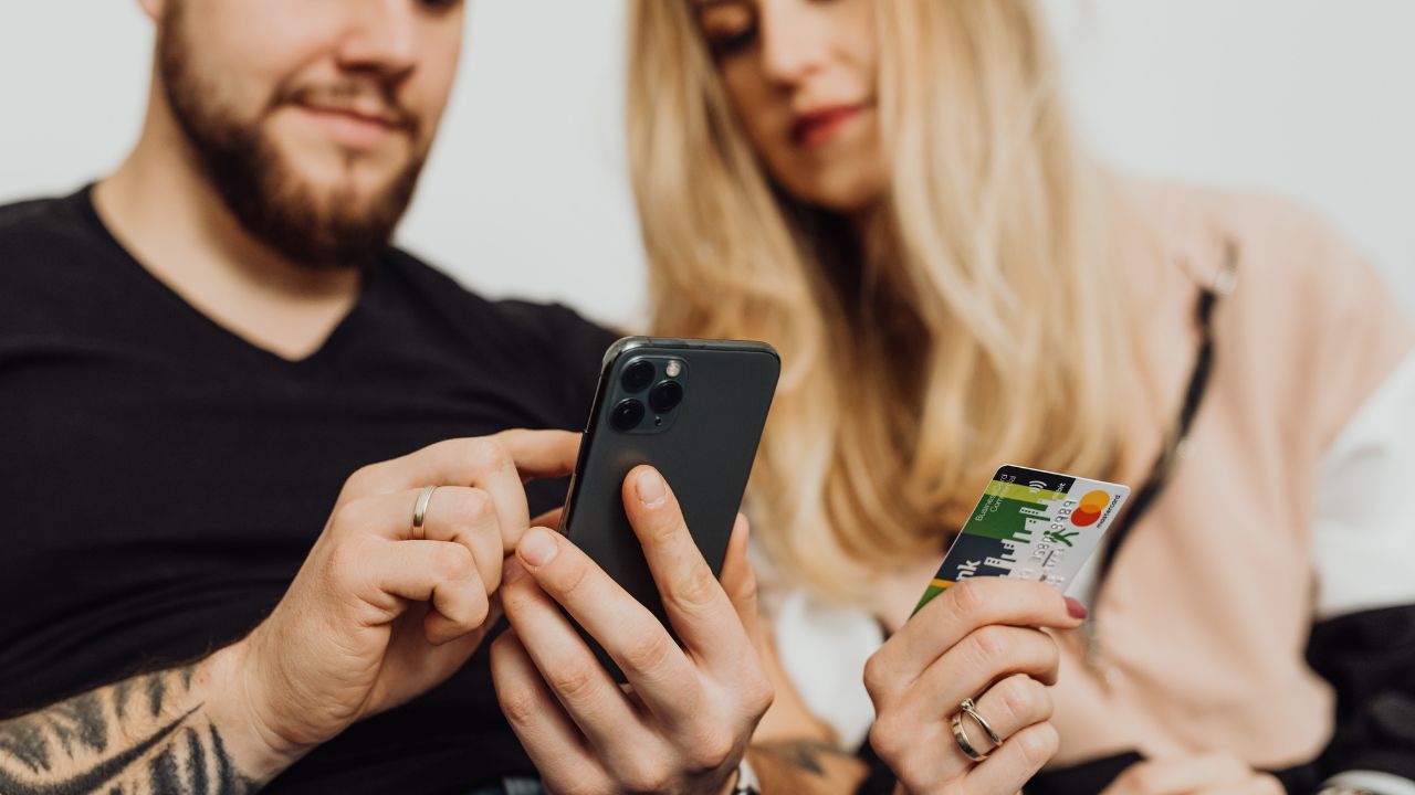 Zwei Personen (Teilansicht) bei einem Online-Bezahlvorgang: Eine Person hält ein schwarzes Smartphone und tippt darauf, die andere Person hält eine Mastercard-Kreditkarte.