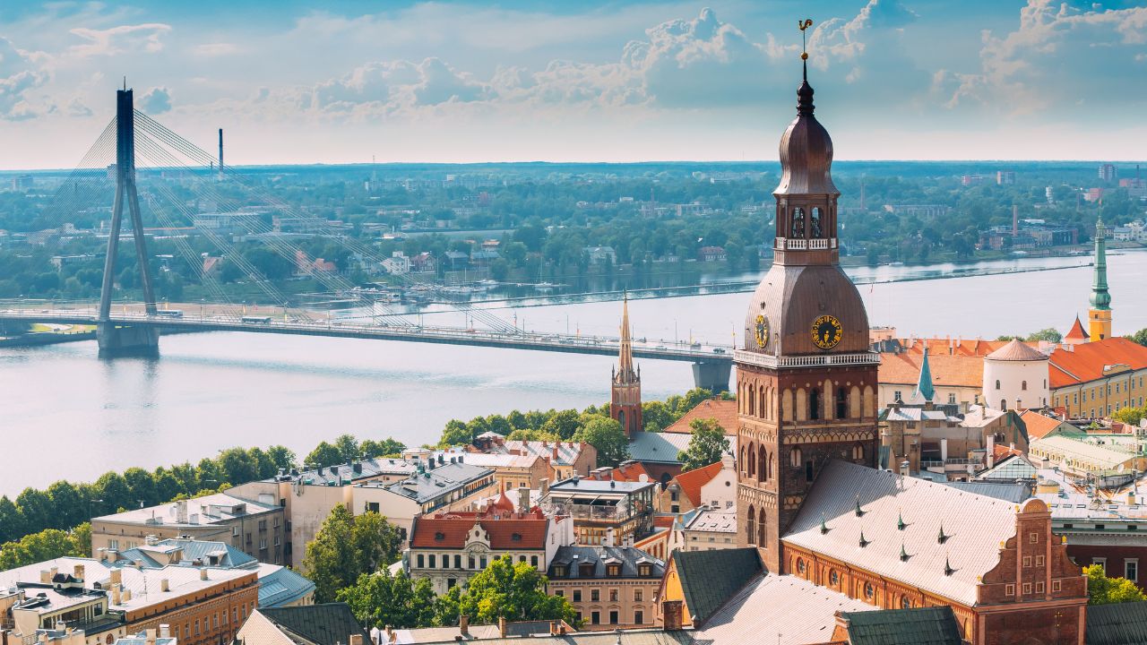 Panoramablick über die Altstadt von Riga, Lettland: Im Vordergrund dominiert der historische, rote Backsteinturm der Petrikirche mit seiner Zwiebelkuppel und den goldenen Uhren. Im Hintergrund überspannt die moderne Vansu-Schrägseilbrücke den breiten Fluss Daugava unter einem wolkenverhangenen Himmel.