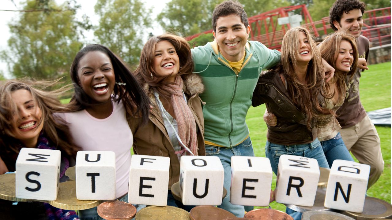 Eine Gruppe von sieben jungen, lachenden Erwachsenen unterschiedlicher Herkunft steht im Freien eng beieinander und blickt fröhlich in die Kamera. Im Vordergrund sind große weiße Buchstabenwürfel aufgestellt, die das Wort 'STEUERN' bilden. Unter den Würfeln liegen Gold- und Kupfermünzen. Der Hintergrund zeigt eine grüne Wiese und eine rote Stahlkonstruktion.