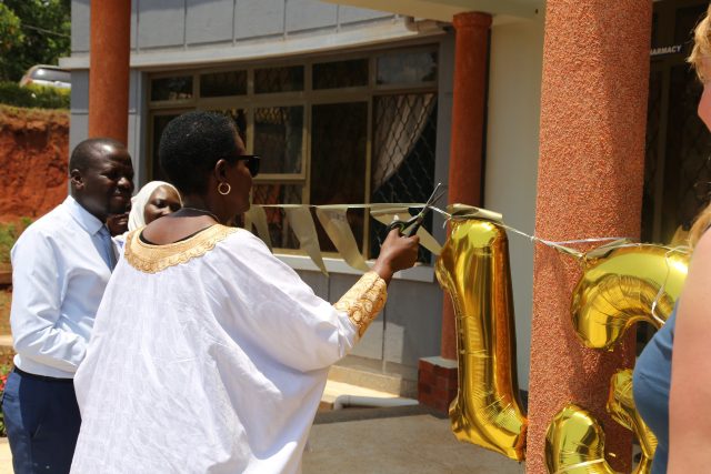 Eine Frau in einem weißen Gewand mit goldener Verzierung durchschneidet mit einer Schere ein goldenes Band bei einer Eröffnungszeremonie vor einem Gebäude, während andere Personen zusehen und goldene Zahlenballons (13) sichtbar sind.