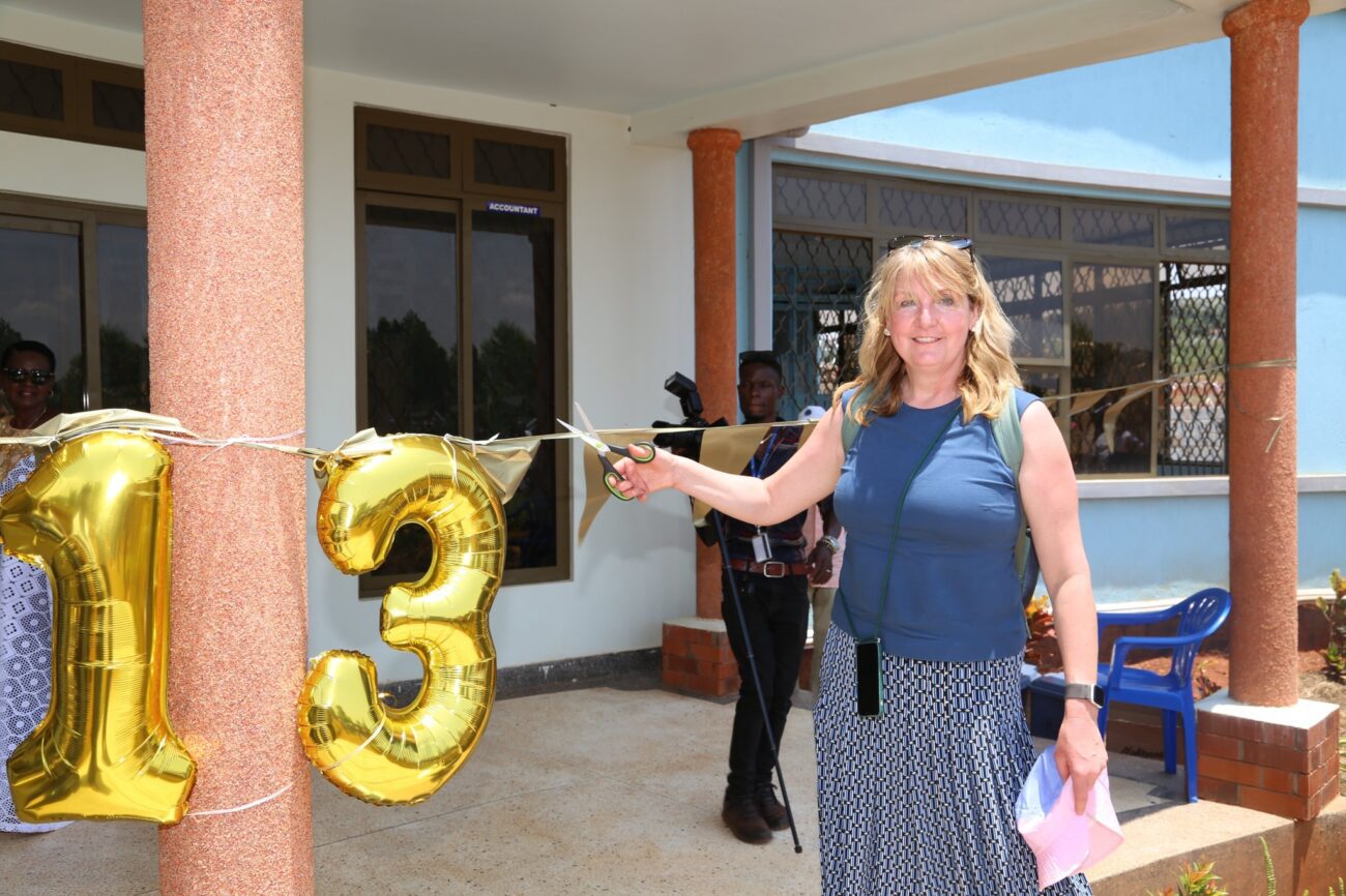 Eine lächelnde Frau mit blonden Haaren durchschneidet mit einer Schere ein Band vor einem Gebäude. Neben ihr hängen goldene Luftballons, die die Zahl 13 bilden.