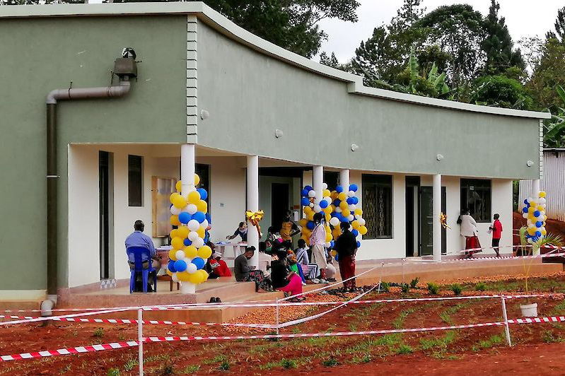Außenansicht eines einstöckigen Gebäudes mit geschwungener, grüner Fassade und überdachter Veranda. Gelb-blaue Luftballons schmücken die Eingangssäulen, während mehrere Menschen auf Bänken sitzen und sich unterhalten. Rot-weißes Absperrband umrahmt das frisch angelegte Gelände im Vordergrund.
