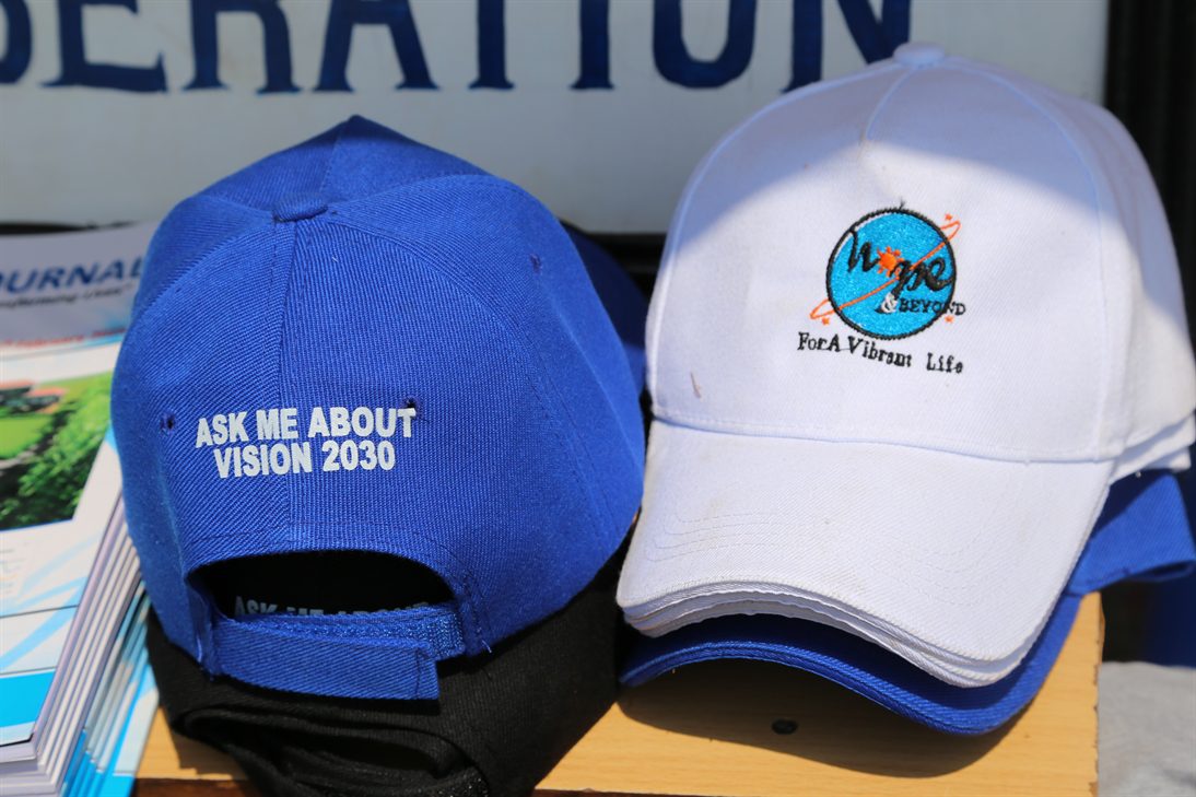 Rückansicht einer blauen Baseballkappe mit der weißen Aufschrift 'ASK ME ABOUT VISION 2030' neben einem Stapel weißer Baseballkappen mit einem Logo mit der Aufschrift 'Hope & Beyond For A Vibrant Life'.