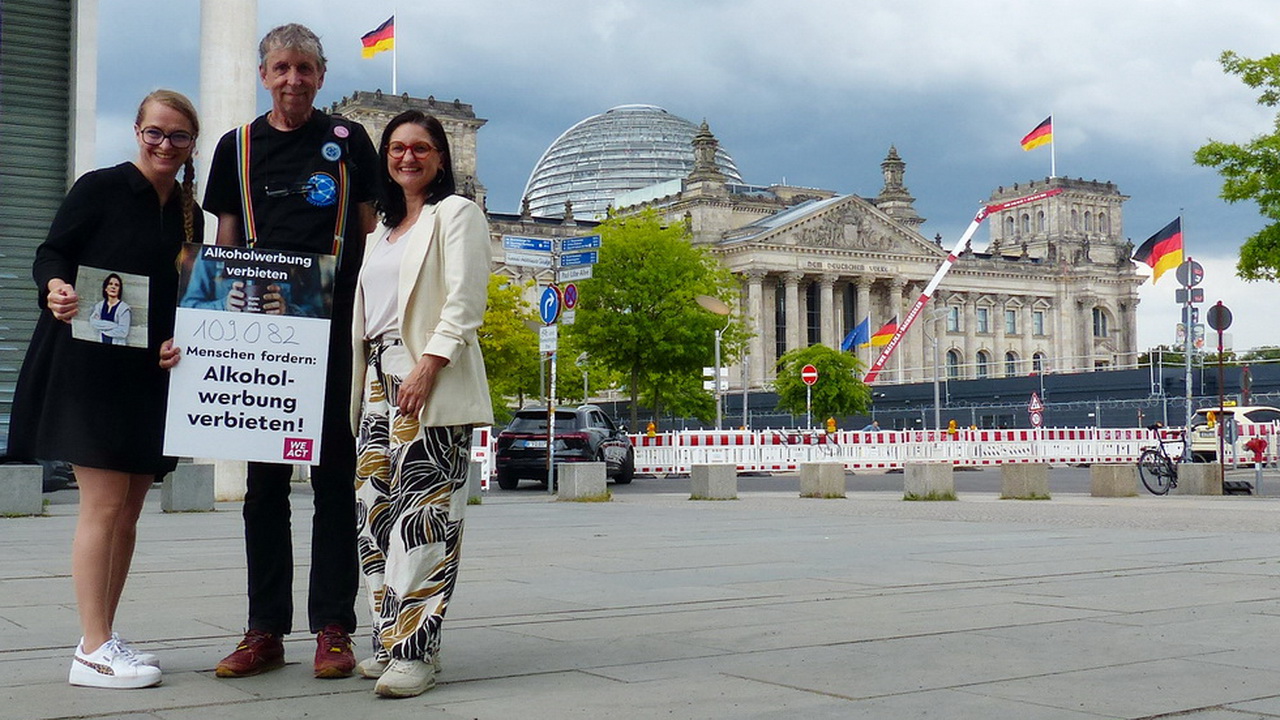 Drei Personen stehen an der Ecke des Paul-Löbe-Hauses in Berlin mit einem Plakat, auf dem steht, dass 109082 Menschen fordern, Alkoholwerbung zu verbieten. Im Hintergrund ist das Gebäude des Bundestages zu sehen.