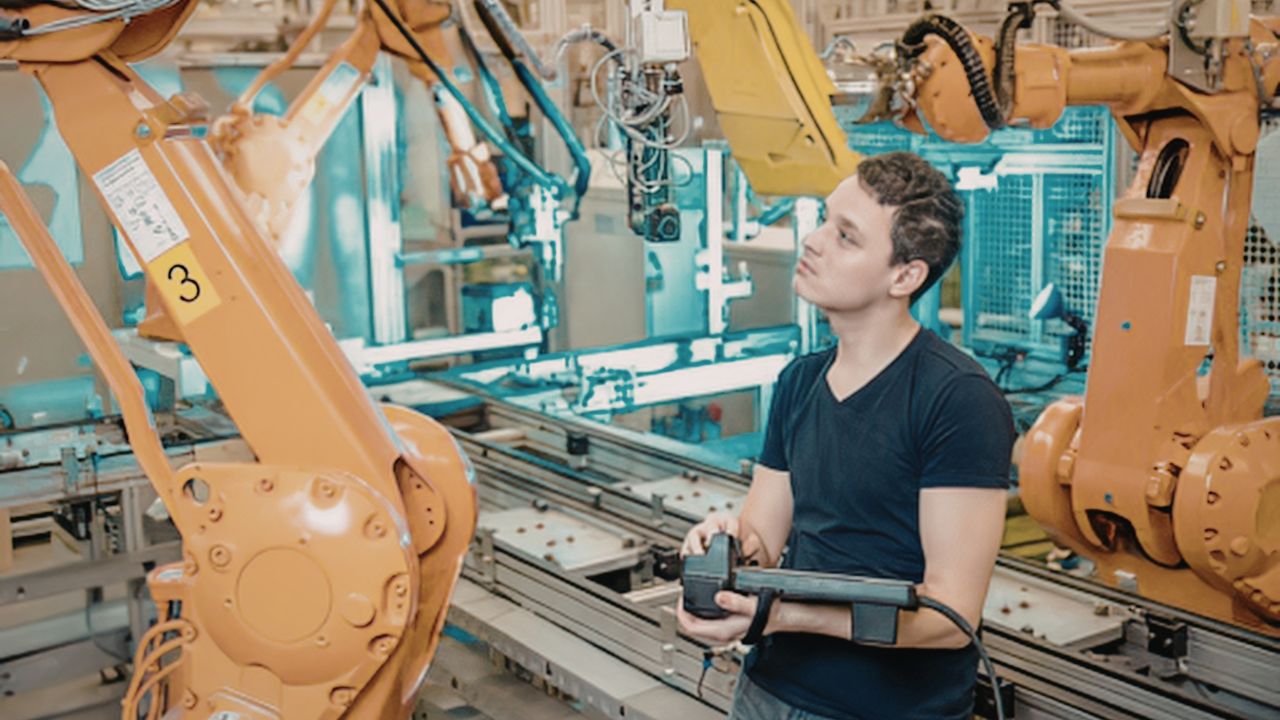 Junger Mann in dunklem T-Shirt bedient ein Handsteuergerät zur Steuerung von orangefarbenen Roboterarmen in einer hell erleuchteten, automatisierten Produktionshalle mit Förderbändern im Hintergrund.