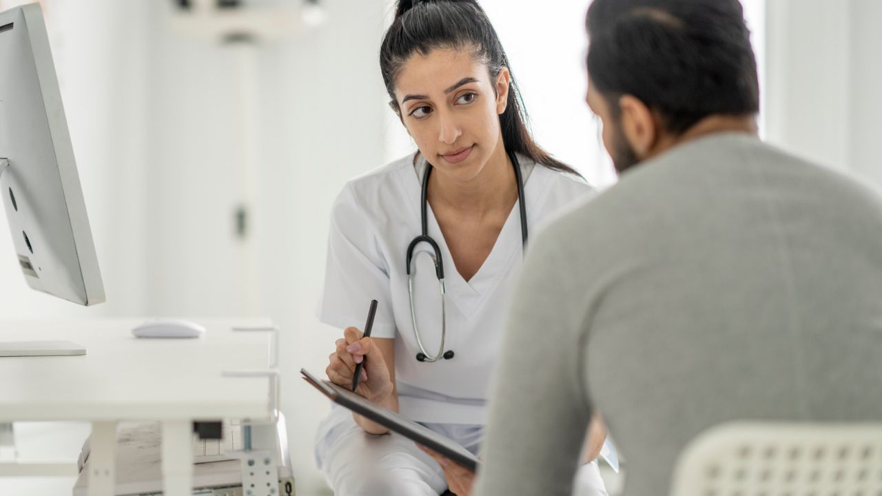 Ärztin in weißer Praxiskleidung mit Stethoskop hört einem Patienten im grauen Pullover aufmerksam zu und hält Stift und Tablet in den Händen, in einem hellen Behandlungszimmer.