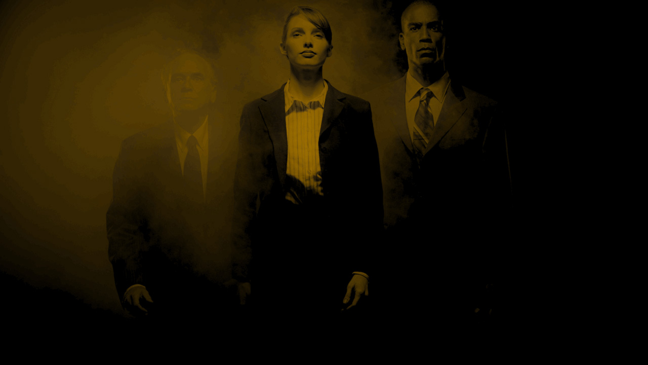 Drei Personen in formeller Kleidung stehen nebeneinander vor einem dunklen, gelblich getönten und von Nebel umgebenen Hintergrund. In der Mitte eine Frau mit kurzen Haaren, flankiert von zwei Männern in Anzügen, alle mit ernstem Gesichtsausdruck.