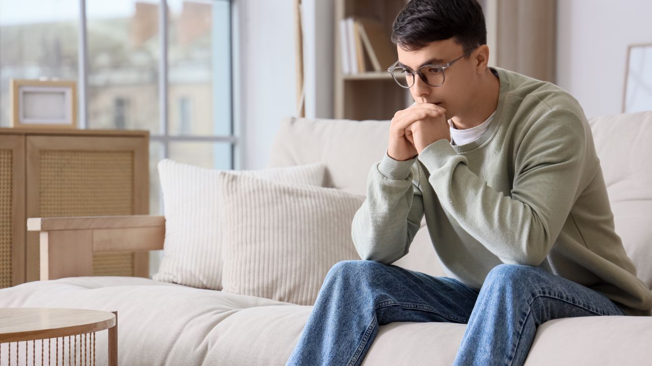Junger Mann mit grauer Brille, hellgrünem Pullover und blauen Jeans sitzt sorgenvoll auf einem beigen Stoffsofa, er hat die Hände vor dem Mund gefaltet und blickt nach unten. Die helle Wohnung ist im skandinavischen Einrichtungsstil mit Holzmöbeln und Rattan-Elementen gestaltet.
