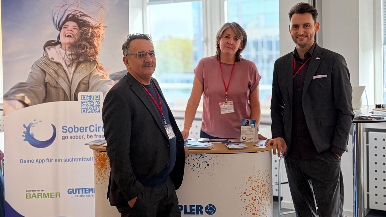 Drei Personen stehen an einem Messestand der App SoberCircle: ein Mann mit Brille und dunklem Anzug, eine Frau mit rosa T-Shirt und ein lächelnder Mann im dunklen Anzug. Im Hintergrund ein Roll-up-Banner mit dem Slogan ‚go sober, be free' und einer lachenden Frau sowie ein Prospekttisch mit Infomaterial.