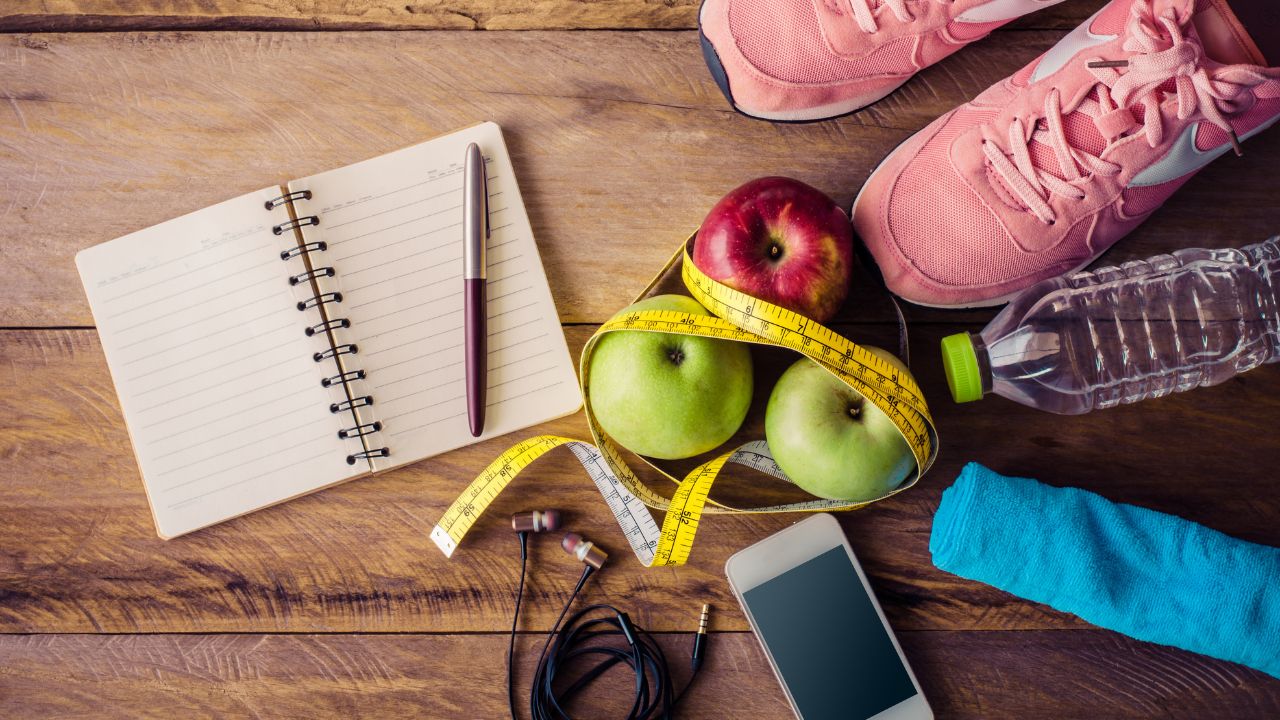 Fitnesszubehör auf Holzboden: pinke Laufschuhe, Äpfel mit Maßband, Wasserflasche, Handtuch, Notizbuch, Smartphone und Kopfhörer.