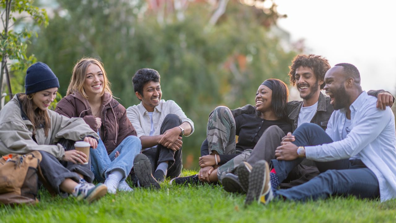 Eine kleine Gruppe junger Erwachsener sitzt gemeinsam im Gras und verbringt Zeit miteinander. Sie sind alle leger in herbstlicher Kleidung angezogen und lachen, während sie die frische Luft und die gemeinsame Zeit genießen.