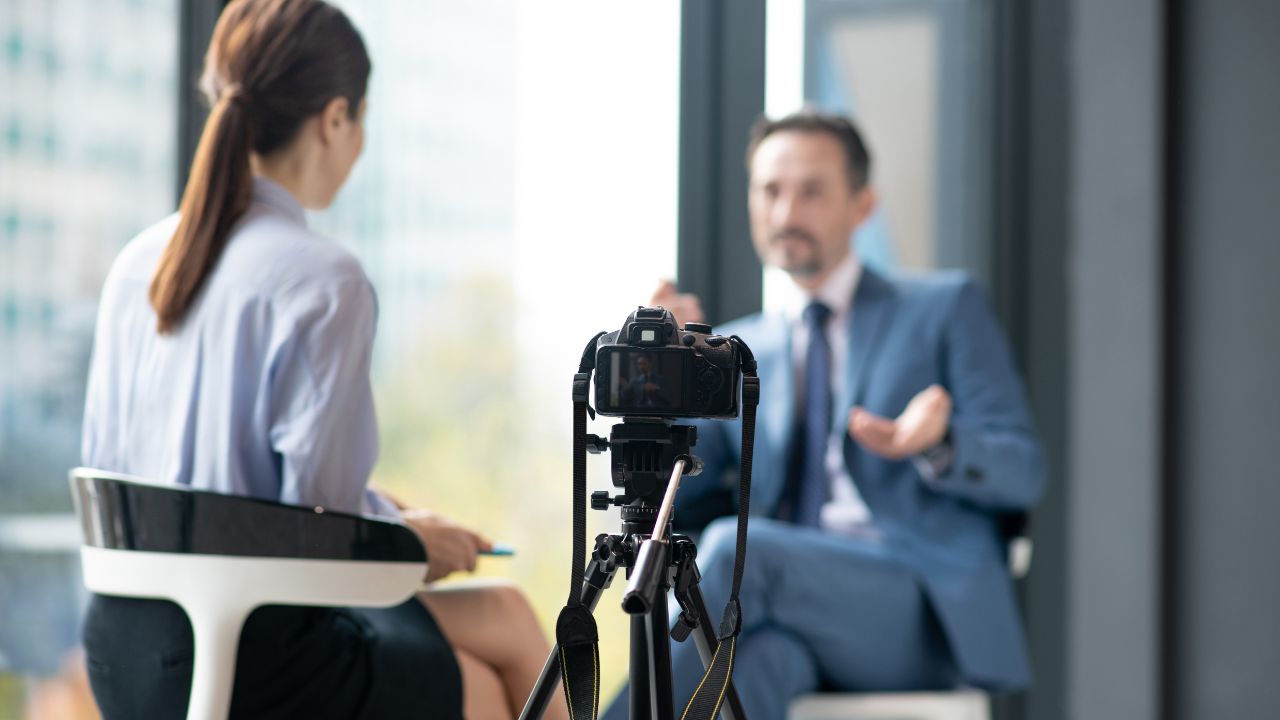 Eine Kamera auf einem Stativ im Vordergrund, scharf gestellt. Im unscharfen Hintergrund führt eine Frau mit Pferdeschwanz ein Interview mit einem Mann in einem hellblauen Anzug in einem hellen Büro mit großen Fenstern.