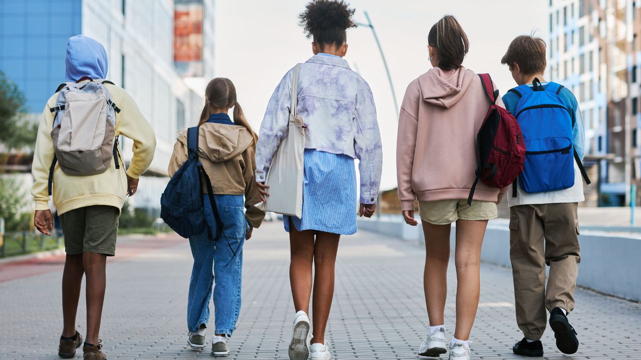 Rückansicht von jungen Schüler*innen mit Rucksäcken, die eine Straße entlanggehen.