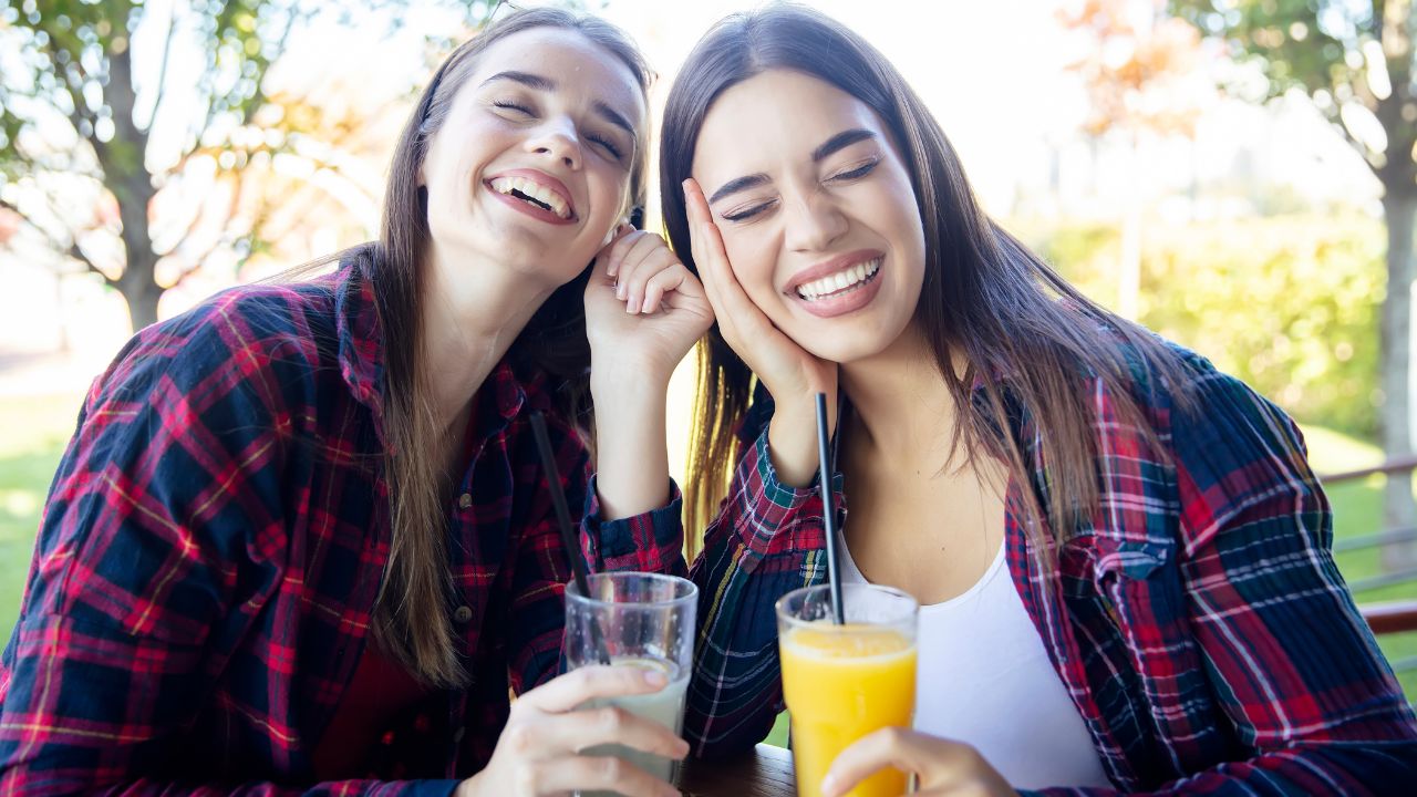 Zwei lachende Freundinnen in rot-blau karierten Hemden sitzen an einem sonnigen Tag im Freien an einem Tisch, lehnen die Köpfe aneinander und halten Gläser mit Orangensaft und Limonade mit Strohhalmen in den Händen; im Hintergrund unscharfe Bäume.
