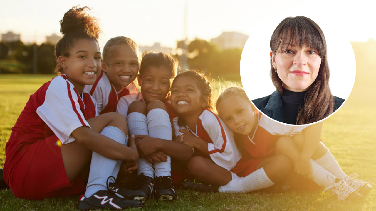 Gruppenfoto von fünf fröhlichen Mädchen in rot-weißen Fußballtrikots auf einem Spielfeld im Abendlicht. Die Kinder unterschiedlicher Herkunft sitzen dicht beieinander auf dem Rasen. Oben rechts freigestellt im Kreis: Kopfporträt von Linda de Keyser mit dunklem Blazer und schwarzem Rollkragenpullover.