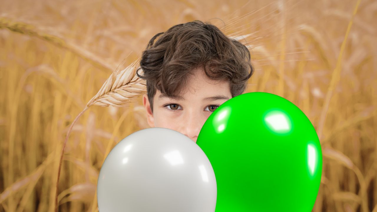 Nahaufnahme eines Jungen mit braunen Locken, der hinter zwei glänzenden Ballons in Weiß und leuchtendem Grün hervorblickt. Im Hintergrund ist ein unscharfes, goldenes Weizenfeld zu sehen.