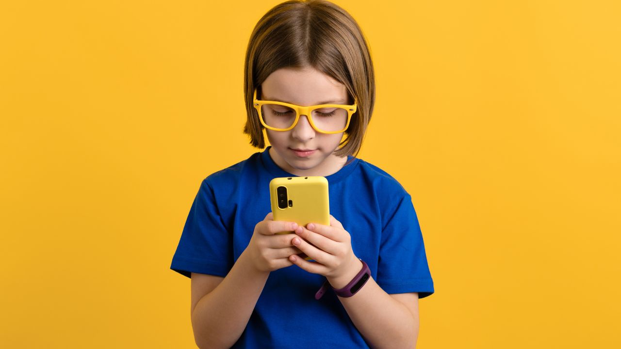 Kind mit gelber Brille und blauem T-Shirt betrachtet ein gelbes Smartphone vor gelbem Hintergrund.